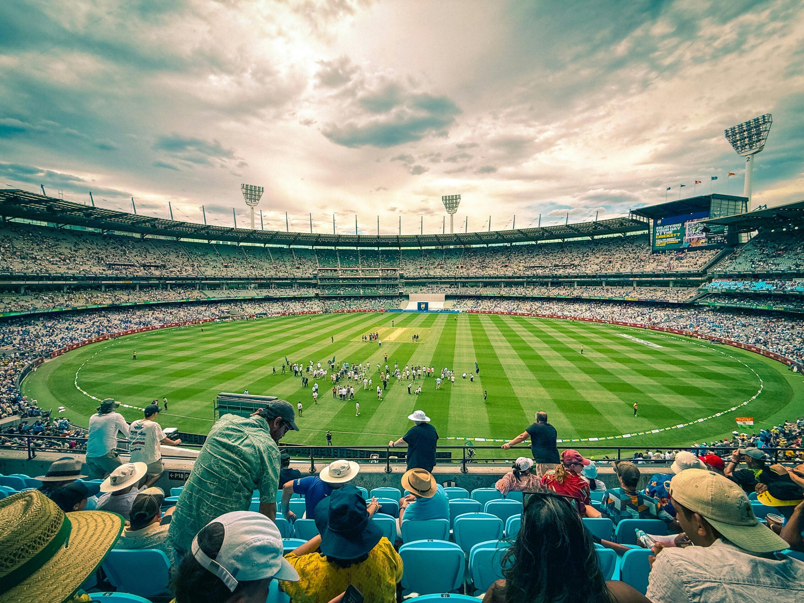 Home A lively cricket match at Melbourne Cricket Ground with a colorful crowd under a dynamic sky.